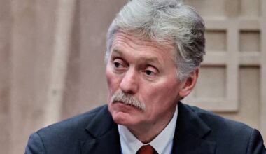 En mann i dress sitter ved et bord under et møte. Han har grått hår og bærer en blå skjorte med slips. I bakgrunnen er en utydelig struktur som gir en avslappet atmosfære. Mannen ser fokusert ut mens han deltar i diskusjonen.