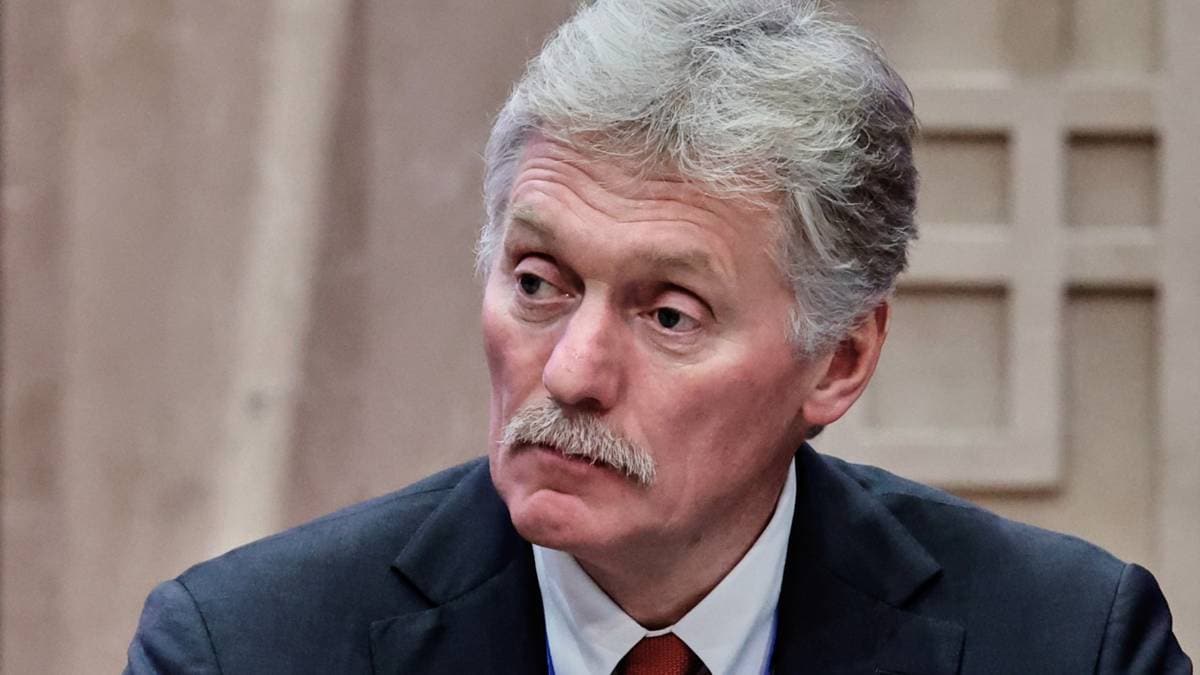 En mann i dress sitter ved et bord under et møte. Han har grått hår og bærer en blå skjorte med slips. I bakgrunnen er en utydelig struktur som gir en avslappet atmosfære. Mannen ser fokusert ut mens han deltar i diskusjonen.