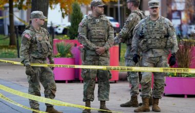 Fire soldater i militæruniformer står sammen ved en avsperring med gult politibånd. De er plassert i et urbant miljø med trær og blomsterkasser i bakgrunnen. En politibil er synlig i avstand. Soldatene ser ut til å overvåke området.