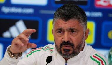 En mann i en hvit jakke sitter bak en mikrofon under en pressekonferanse. Han gestikulerer med hånden mens han snakker. Bakgrunnen inneholder flere logoer og fargerike elementer. Det er tydelig at mannen diskuteres i en profesjonell setting.