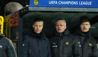 Fire personer står på en trenerbenk under en UEFA Champions League-kamp. En av dem er iført en mørk treningsjakke. I bakgrunnen er det tilskuere og en stor skjerm som viser "UEFA CHAMPIONS LEAGUE". Omgivelsene indikerer at bildet er tatt på en fotballstadion.