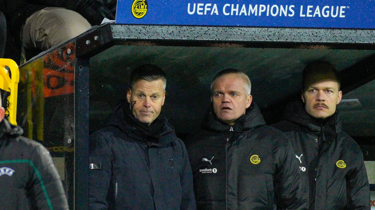 Fire personer står på en trenerbenk under en UEFA Champions League-kamp. En av dem er iført en mørk treningsjakke. I bakgrunnen er det tilskuere og en stor skjerm som viser "UEFA CHAMPIONS LEAGUE". Omgivelsene indikerer at bildet er tatt på en fotballstadion.