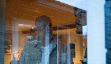 To personer står ved et vindu, med en innendørs setting synlig i bakgrunnen. Den ene personen har på seg en lys jakke og ser ut av vinduet. Møbler og belysning kan sees i rommet bak dem. Lysforholdene indikerer en dyster stemning.