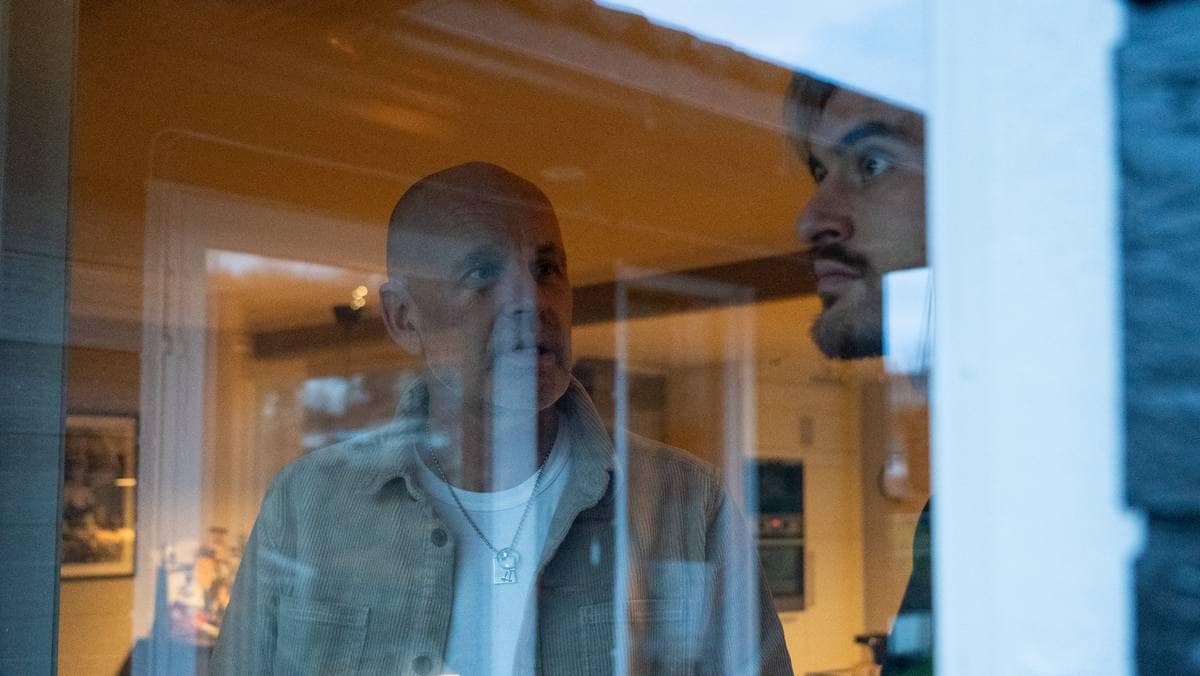 To personer står ved et vindu, med en innendørs setting synlig i bakgrunnen. Den ene personen har på seg en lys jakke og ser ut av vinduet. Møbler og belysning kan sees i rommet bak dem. Lysforholdene indikerer en dyster stemning.