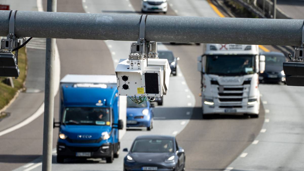 Et overvåkingskamera henger fra en lyktestolpe over en motorvei. Flere kjøretøy, inkludert en lastebil og personbiler, kjører i høyre kjørefelt. Omgivelsene viser en asfaltvei med to kjørefelt og grønt gress på venstre side. Bildeutsnittet indikerer en travel vei med aktiv trafikk.