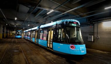 To trikker, en mørk blå og en lyseblå, står i en pittoresk sporvognshal. Den lyseblå trikken har julelys og viser teksten "God jul". Omgivelsene er preget av betongvegger og sporveier. Det er flomlys som belyser området.