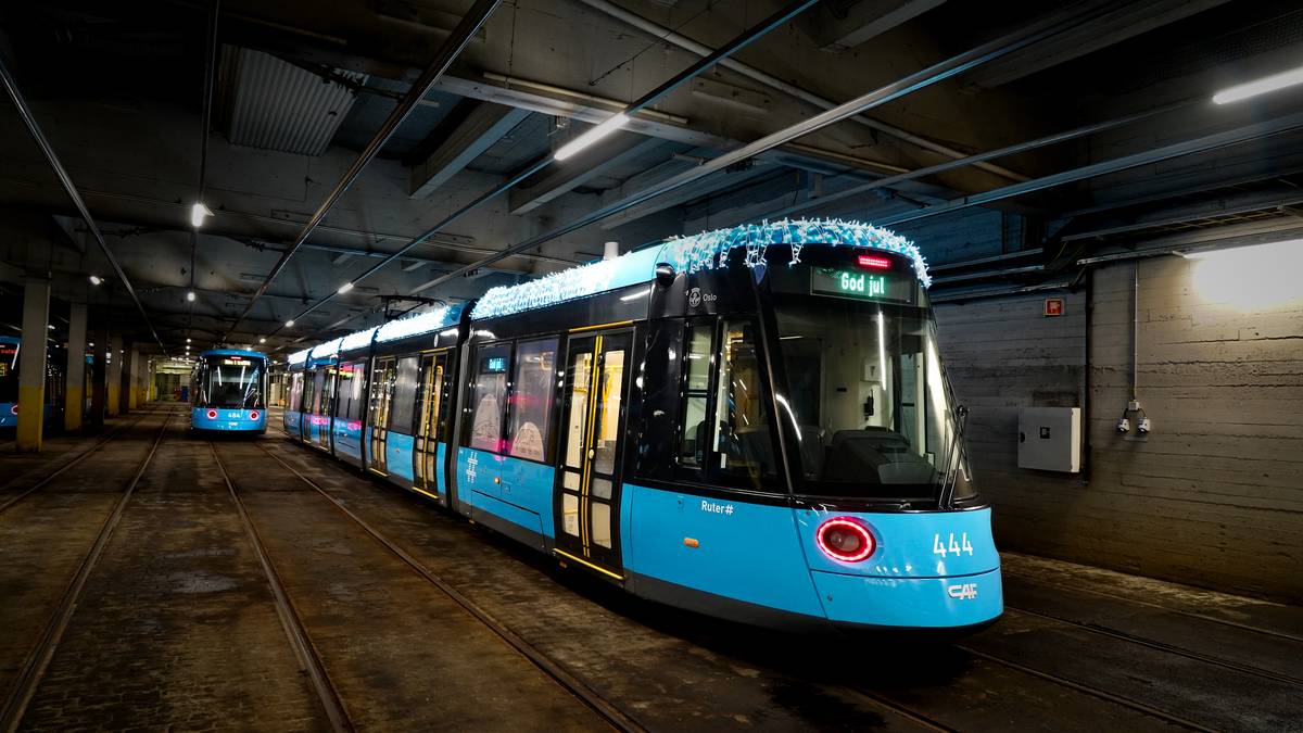 To trikker, en mørk blå og en lyseblå, står i en pittoresk sporvognshal. Den lyseblå trikken har julelys og viser teksten "God jul". Omgivelsene er preget av betongvegger og sporveier. Det er flomlys som belyser området.