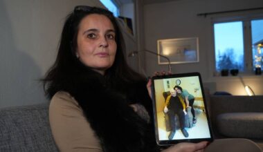 En kvinne sitter på en sofa og holder opp en nettbrett. På skjermen vises to personer, en som sitter i en stol og en som står bak. Omgivelsene er et rom med møbler og et vindu som slipper inn naturlig lys. Det er lysere farger på veggene og interiøret.