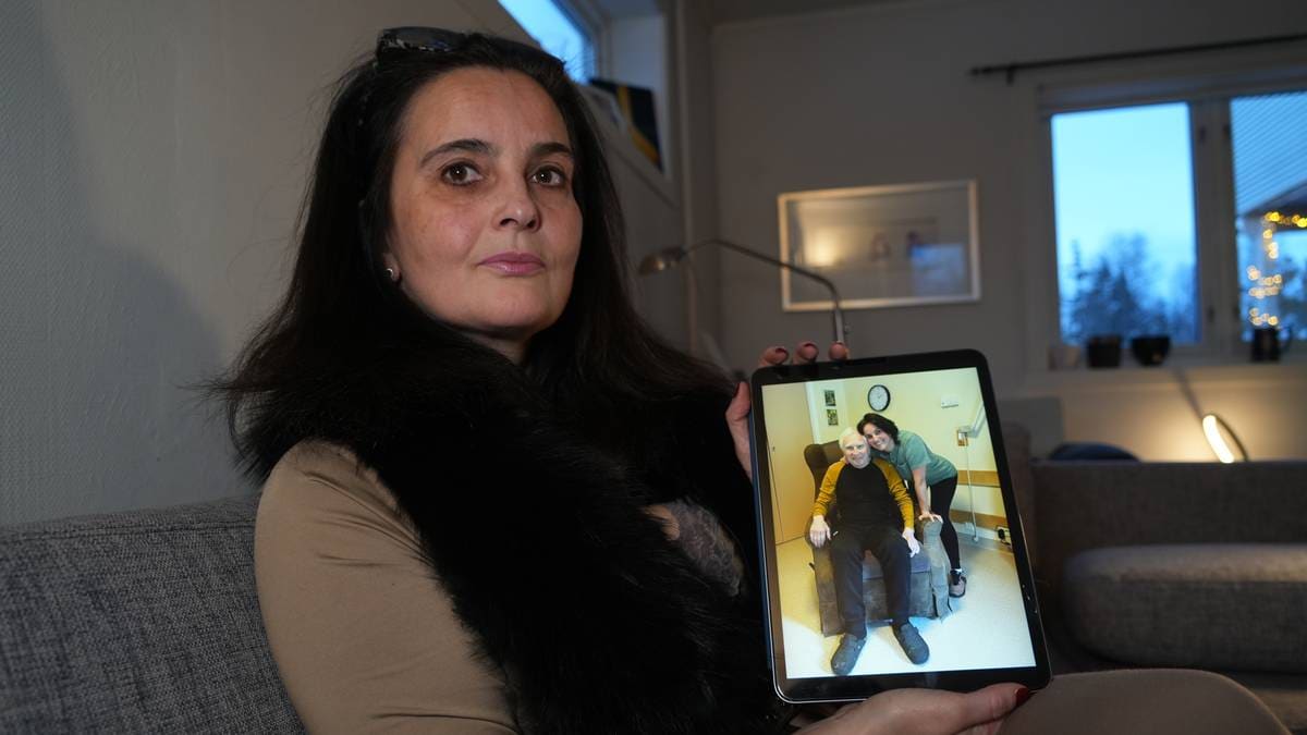 En kvinne sitter på en sofa og holder opp en nettbrett. På skjermen vises to personer, en som sitter i en stol og en som står bak. Omgivelsene er et rom med møbler og et vindu som slipper inn naturlig lys. Det er lysere farger på veggene og interiøret.