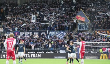 Et fotballag spiller på banen, mens en stor mengde supportere heier i tribunene. Supporterne holder opp skjerf og flagg med klubbens logoer. I forgrunnen står spillere, en i rød drakt og to i mørk drakt, som ser mot hverandre. Omgivelsene viser stadion med tydelig publikum og spilleforhold.