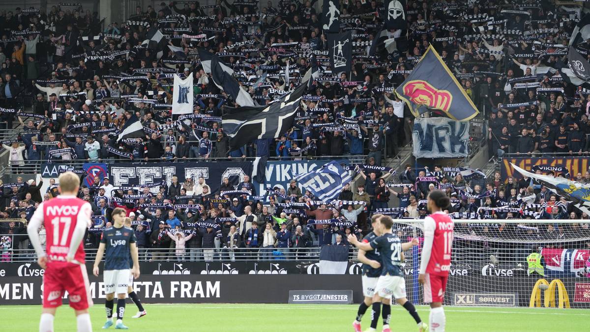 Et fotballag spiller på banen, mens en stor mengde supportere heier i tribunene. Supporterne holder opp skjerf og flagg med klubbens logoer. I forgrunnen står spillere, en i rød drakt og to i mørk drakt, som ser mot hverandre. Omgivelsene viser stadion med tydelig publikum og spilleforhold.