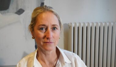 En kvinne sitter foran en radiator, iført en hvit skjorte med smykker. Hun har blond hår og det er et abstrakt maleri i bakgrunnen. Det er et rolig og innendørs miljø. Mikrofonen bort til venstre antyder at hun deltar i et intervju.