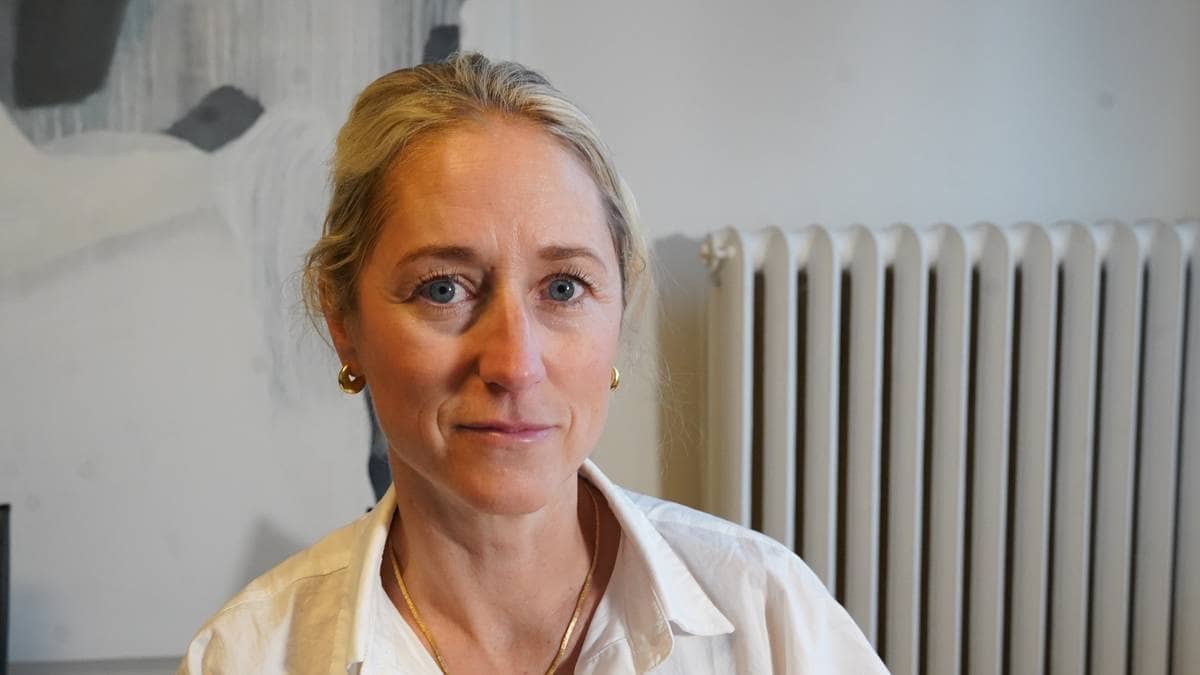 En kvinne sitter foran en radiator, iført en hvit skjorte med smykker. Hun har blond hår og det er et abstrakt maleri i bakgrunnen. Det er et rolig og innendørs miljø. Mikrofonen bort til venstre antyder at hun deltar i et intervju.