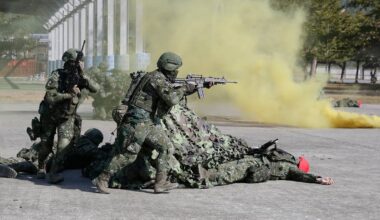 Soldater i kamuflasje deltar i trening med røyk granater. En soldat holder gevær, mens andre tar dekning. Omgivelsene viser et militært treningsområde. Gult røyk legger seg i luften, som bidrar til treningssituasjonen.
