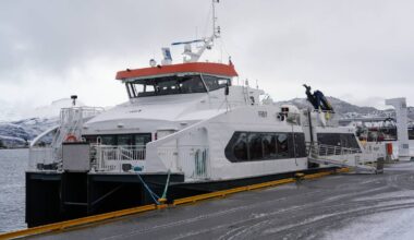 Fergen "Frey" ligger ved kaien i en havn med snødekte fjell i bakgrunnen. Det er grå himmel, og det synes å være kaldt vær. En person er synlig ved fergen, trolig i ferd med å gjøre forberedelser. Kaien er utstyrt med guldkledde stropper og utstyr for lasting av varer.