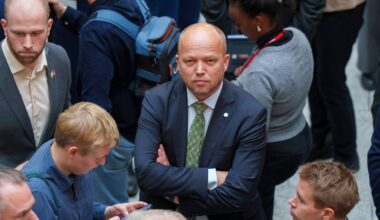 Flere personer står tett sammen, med en mann i dress i fokus. Han har armene krysset og virker ettertenksom. I nærheten ser vi yngre mennesker som bruker mobiltelefoner. Omgivelsene er en folkemengde, trolig i en offentlig setting.