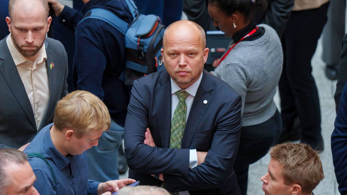 Flere personer står tett sammen, med en mann i dress i fokus. Han har armene krysset og virker ettertenksom. I nærheten ser vi yngre mennesker som bruker mobiltelefoner. Omgivelsene er en folkemengde, trolig i en offentlig setting.