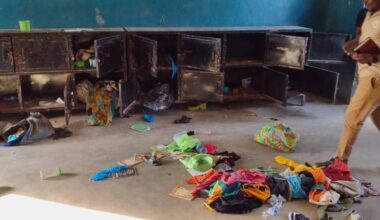 Rottede klær ligger spredt på gulvet i et rom med blå vegger. En mann i lys skjorte og bukser står ved skapet, som har åpne dører. Han holder en notatbok og ser mot skapene. Rommet virker kaotisk, med flere gjenstander og klær liggende rundt.