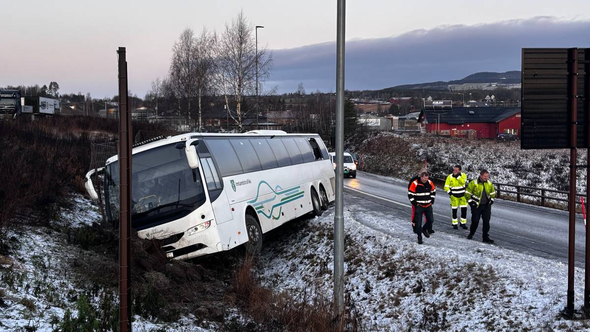 Et passasjerknutebuss står delvis utfor veien i en snødekt skråning. Flere personer i arbeidstøy, inkludert redningsmannskap, inspiserer hendelsen. I bakgrunnen er det en tydelig vei med biler og noen bygninger. Omgivelsene viser en kald og vinterlig atmosfære.