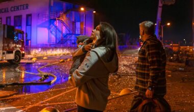 En kvinne holder et barn mens hun ser på brannbilen ved et medisinsk senter. En mann i rutete skjorte står ved siden av henne. Området er opplyst av blåaktige lys fra brannbilen, og bakken er dekket av ødelagte gjenstander. Det er spor av vannslanger og rusk fra en muligens tidligere brann.
