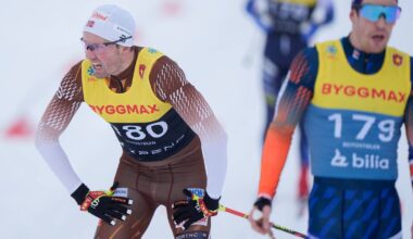 To personer i skiklær står på snøen. Den ene personen, iført en gul startnummertrøye med nummer 180, bøyer seg fremover og ser ut til å ta seg en pause. Den andre personen, med startnummer 179, står i bakgrunnen og ser mot konkurransen. Omgivelsene er preget av snødekte bakker og skistaver.