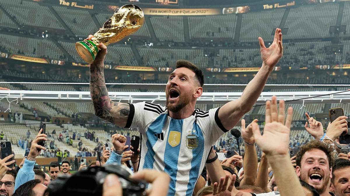 En fotballspiller i Argentinas drakt hever FIFA verdensmesterskapstroféet. Han er omringet av feirende supportere. Arenaen har en stor kapasitet, og mange tilskuere er til stede. Stemningen er festlig, med folk som tar bilder og jubler.