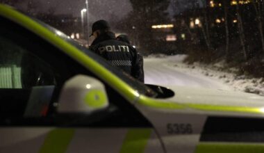 To politibetjenter står nær en bil i snøfylt gate om natten. Den ene betjenten har på seg en jakke med teksten "POLITI". Gatebelysning og snøfall bidrar til atmosfæren. En del av en politibil med karakteristiske farger er synlig i forgrunnen.