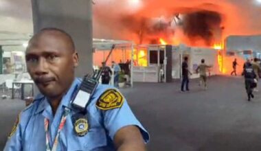 En varemesse opplever en brann. Røyk og flammer er synlige i bakgrunnen, og personer evakuerer området. En sikkerhetsvakt står foran kameraet, iført uniform. Omgivelsene er preget av panikk og hastverk blant de tilstedeværende.