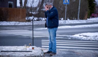 En mann står ved kanten av veien, med hendene i ansiktet. Foran ham er det plassert flere lys på snøen. Området er preget av vinter med snø på bakken og en asfaltvei bak mannen. Det finnes også et trafikkskilt og et gjerde i bakgrunnen.