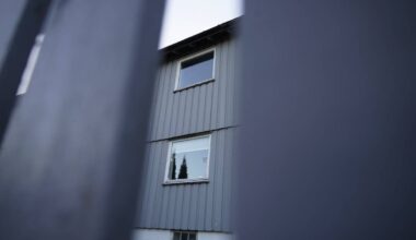 Bildet viser et grått hus sett gjennom en gjerde. To vinduer er synlige, med trær i bakgrunnen. Omgivelsene fremstår som liberale og nabolaget ser rolig ut. Lyset er dempet, trolig sent på ettermiddagen.