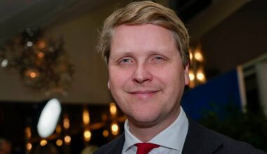 En mann iført dress og rød slips står i en social setting med dempet belysning. Det er utydelig bakgrunn med dekorative elementer. Stemningen ser ut til å være uformell, muligens en sammenkomst. Lysene i rommet bidrar til en avslappet atmosfære.