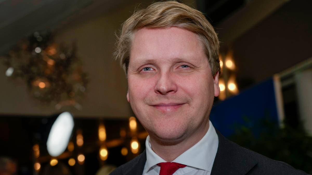 En mann iført dress og rød slips står i en social setting med dempet belysning. Det er utydelig bakgrunn med dekorative elementer. Stemningen ser ut til å være uformell, muligens en sammenkomst. Lysene i rommet bidrar til en avslappet atmosfære.