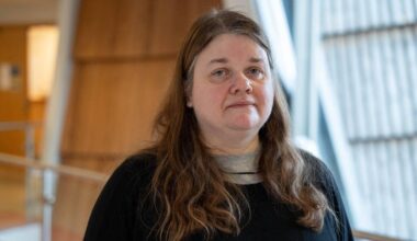 En kvinne med langt, brunt hår står foran en moderne bygning. Hun er kledd i en svart genser og en grå t-skjorte. Omgivelsene har glassvegger og treverk, som gir et åpent inntrykk. Det er naturlig lys i rommet.