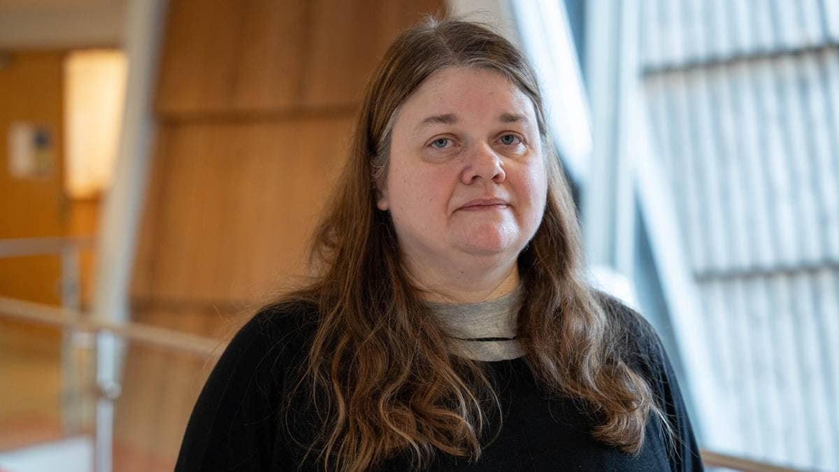 En kvinne med langt, brunt hår står foran en moderne bygning. Hun er kledd i en svart genser og en grå t-skjorte. Omgivelsene har glassvegger og treverk, som gir et åpent inntrykk. Det er naturlig lys i rommet.