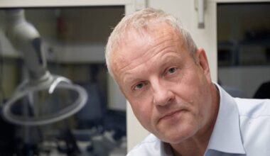 En mann sitter ved et bord med teknisk utstyr i bakgrunnen. Han har grått hår og bærer en lyseblå skjorte. Det er et arbeidsmiljø med flere apparater synlige rundt ham. Bordet er dekket med diverse gjenstander og kabler.