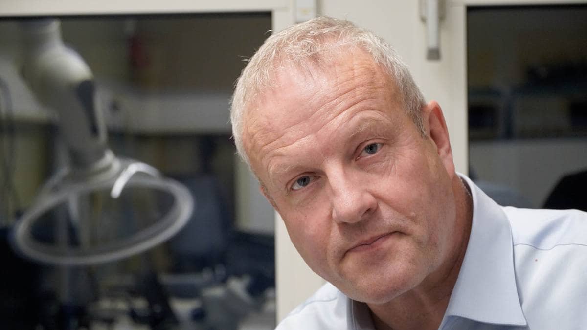 En mann sitter ved et bord med teknisk utstyr i bakgrunnen. Han har grått hår og bærer en lyseblå skjorte. Det er et arbeidsmiljø med flere apparater synlige rundt ham. Bordet er dekket med diverse gjenstander og kabler.