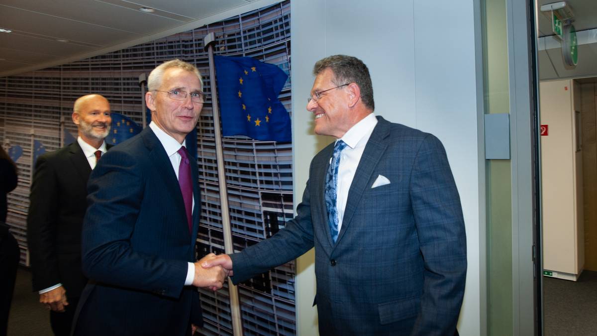 To personer håndhilser i en moderne kontorinnstilling. Europeiske flagg er synlige i bakgrunnen. Det er flere personer i nærheten som observerer situasjonen. Miljøet gir inntrykk av en formell møteplass.