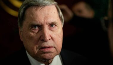 Jurij Usjakov snakker med journalister under en pressekonferanse. Kamerautstyr og mikrofoner er til stede. Bakgrunnen er uklar, men antyder et formelt miljø. Lyskilder skaper en dempet atmosfære i rommet.