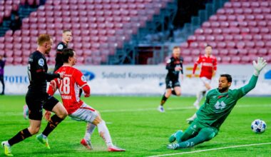 En spiller fra laget i rødt, med draktnummer 28, er i ferd med å skyte ballen mot mål. Målmannen i grønn drakt strekker seg for å redde. I bakgrunnen er flere spillere fra begge lag synlige. Omgivelsene inkluderer et fotballstadion med tribuner og flomlys.