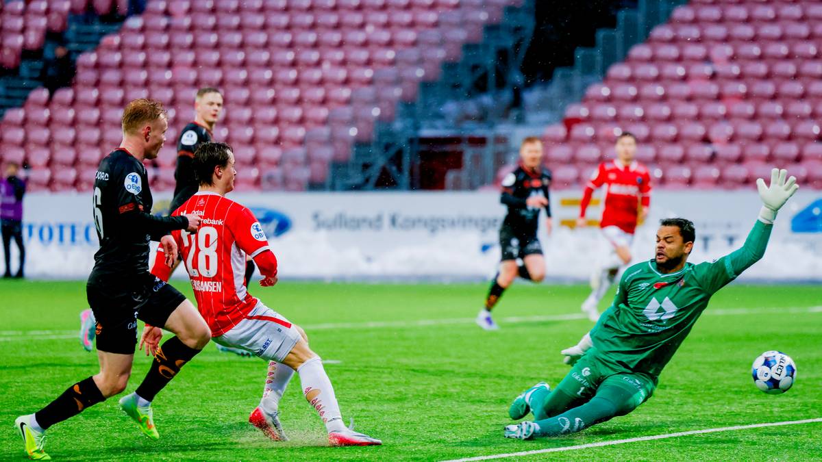 En spiller fra laget i rødt, med draktnummer 28, er i ferd med å skyte ballen mot mål. Målmannen i grønn drakt strekker seg for å redde. I bakgrunnen er flere spillere fra begge lag synlige. Omgivelsene inkluderer et fotballstadion med tribuner og flomlys.