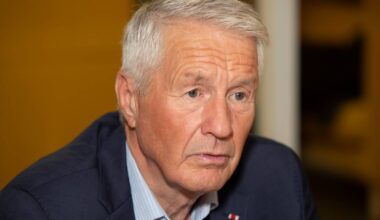 En eldre mann sitter med hodet skrått og ser mot venstre. Han har grått hår og bære en mørk dressjakke over en skjorte med prikker. Bakgrunnen er uskarp, men gir inntrykk av en innendørs setting. Det er en generell atmosfære av samtale eller diskusjon.