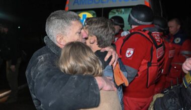 To personer omfavner hverandre foran en ambulanse. Redningsarbeidere i uniformer er synlige i bakgrunnen. Omgivelsene er dålige opplyst, noe som gir en trist stemning. Bildet kan indikere en nødssituasjon eller redningsoperasjon.