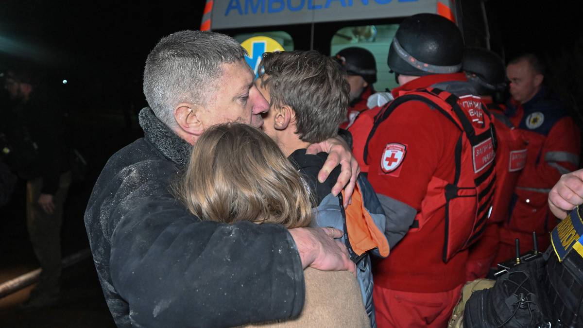 To personer omfavner hverandre foran en ambulanse. Redningsarbeidere i uniformer er synlige i bakgrunnen. Omgivelsene er dålige opplyst, noe som gir en trist stemning. Bildet kan indikere en nødssituasjon eller redningsoperasjon.