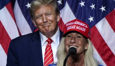 En mann i dress med slips står bak en kvinne iført en blå topp og en rød caps. Kvinnen taler fra en mikrofon. Bakgrunnen består av amerikanske flagg. Begge ser mot publikum under et arrangement.