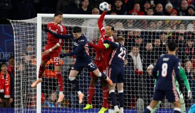 To spillere fra hvert lag kjemper om ballen i en fotballkamp, bakgrunnen viser et fullt stadion. Den ene spilleren i rødt hopper opp for å blokkere et skudd. De øvrige spillerne er i bevegelse, fokusert på spillet. Dommeren og tilskuere i bakgrunnen bidrar til settingen.