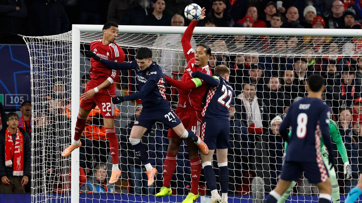 To spillere fra hvert lag kjemper om ballen i en fotballkamp, bakgrunnen viser et fullt stadion. Den ene spilleren i rødt hopper opp for å blokkere et skudd. De øvrige spillerne er i bevegelse, fokusert på spillet. Dommeren og tilskuere i bakgrunnen bidrar til settingen.