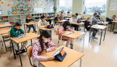 Elever sitter ved pultene i et klasserom, engasjert i lesing. De bruker både bøker og nettbrett. Rommet er lys og fargerikt, med kunstverk og dekorasjoner på veggene. Det er flere vinduer som gir naturlig lys til rommet.