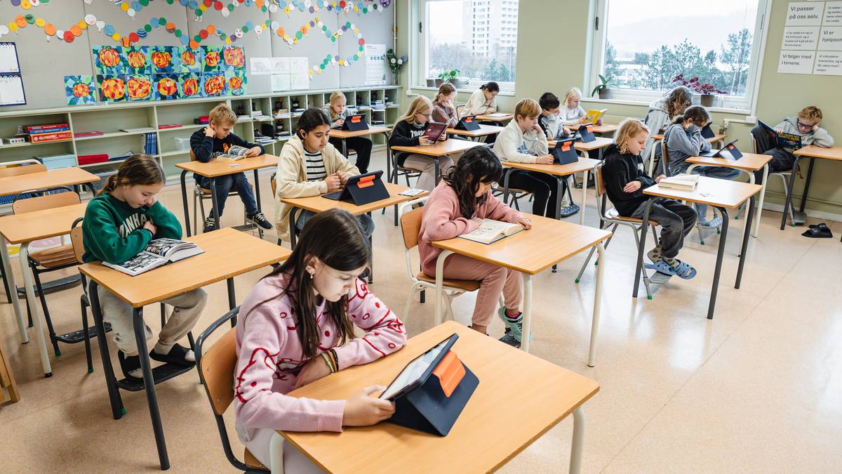 Elever sitter ved pultene i et klasserom, engasjert i lesing. De bruker både bøker og nettbrett. Rommet er lys og fargerikt, med kunstverk og dekorasjoner på veggene. Det er flere vinduer som gir naturlig lys til rommet.