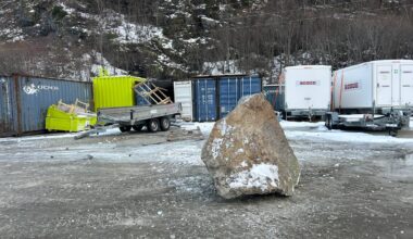 Et stort steinblokk ligger på bakken på en skrotplass. Rundt steinen er det flere fraktcontainere og en tilhenger. Området er dekket av snø, og fjell i bakgrunnen skaper en kald atmosfære. Det er ingen personer synlige i bildet.