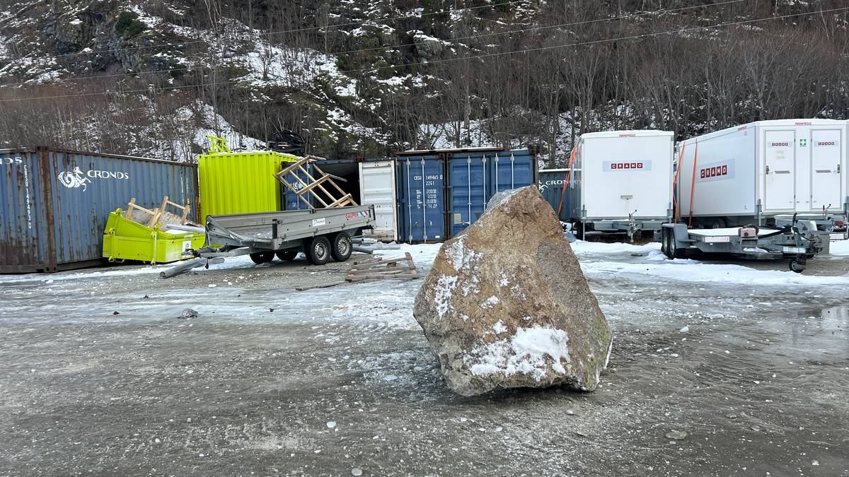 Et stort steinblokk ligger på bakken på en skrotplass. Rundt steinen er det flere fraktcontainere og en tilhenger. Området er dekket av snø, og fjell i bakgrunnen skaper en kald atmosfære. Det er ingen personer synlige i bildet.